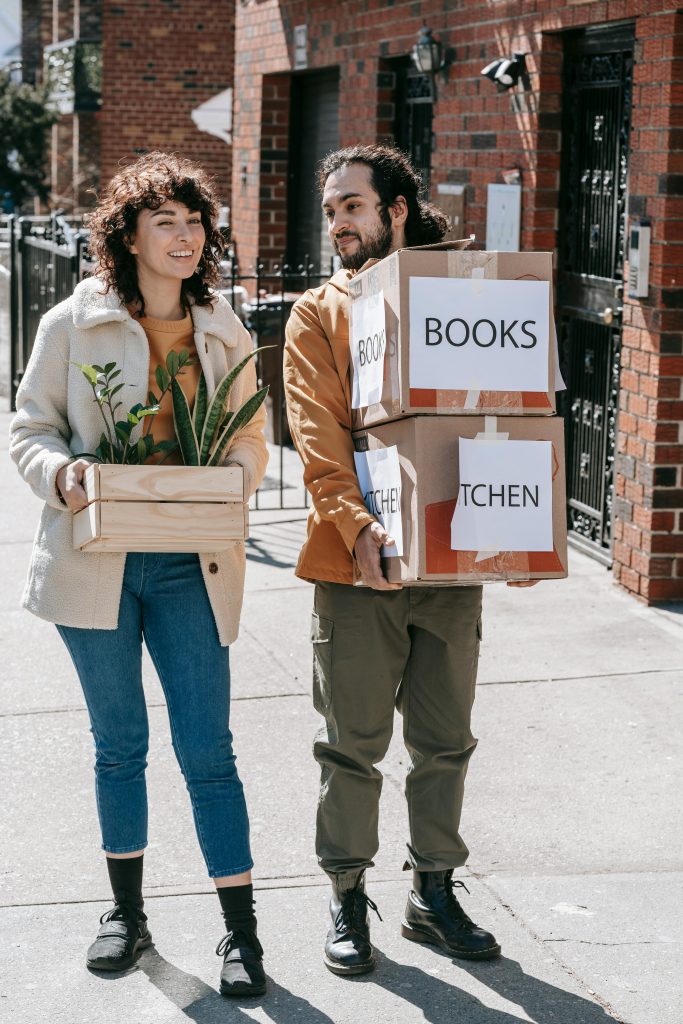 Couple smiling while carrying boxes and plants during urban relocation in daylight.